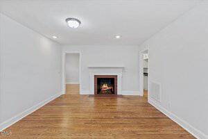 A living room with hardwood floors, white walls, a white mantel fireplace with a fire burning, overhead lights, and doorways leading to adjacent rooms.