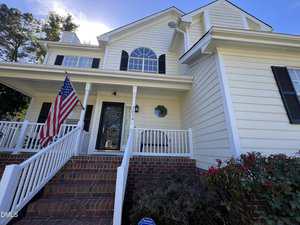 A two-story yellow house with white trim, black shutters, a front porch, brick steps, an American flag on a pole, and a bush in the foreground under a blue sky.
