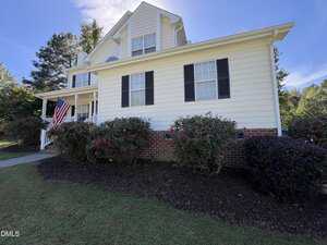 A two-story house with light yellow siding, black shutters, and a brick foundation. Bushes with pink flowers line the front, and an American flag is displayed by the porch. The sky is clear and blue.