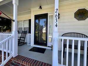 A covered front porch with white railings, a black rocking chair, a black bench, a glass front door with a decorative design, house number 2634, and a stained-glass window on the right wall.