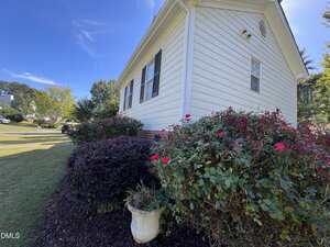 A white house with black shutters is bordered by green shrubs and red flowering bushes. There is a white planter in front of the bushes, and the lawn and neighboring houses are visible under a clear blue sky.