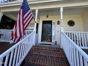 A front porch with brick steps, white railings, and a glass door. An American flag hangs on the left. The house number 2634 is displayed on a post. A small decorative statue sits near the door.