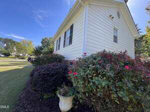 A white house with black shutters is shown from the side, bordered by bushes with red flowers and a white planter in front. The lawn is neatly trimmed and the sky is clear and blue.