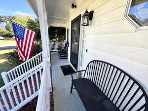 A front porch with a black metal bench, two rocking chairs, a brick step, and an American flag hanging from a white railing. A glass front door and a lantern-style light fixture are visible.