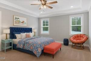 A bedroom with a blue tufted bed, orange patterned bedding, two blue nightstands with lamps, an orange bench, a round orange chair, beige carpet, and two windows with white blinds. A ceiling fan and wall art are visible.