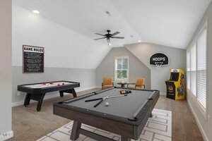 A game room with a pool table, air hockey table, two armchairs with a small table, an arcade machine, a wall-mounted basketball hoop, large windows, and a ceiling fan. A sign with game rules hangs on the wall.
