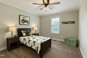 A child's bedroom with a wooden bed, tractor-themed bedding, two bedside tables with lamps, a wall art of a green tractor, a green toy chest, and the word "TRACTOR" on the wall. A window lets in natural light.
