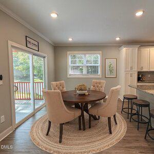 A dining area with a round wooden table, four beige upholstered chairs on a round rug, next to a kitchen with bar stools and granite countertops. Large windows and sliding glass doors provide natural light.