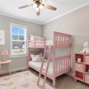 A small bedroom with gray walls features a pink bunk bed, a window with white trim, a ceiling fan, a pink lamp on a round table, cubed storage with stuffed toys, and a heart-patterned rug on the floor.
