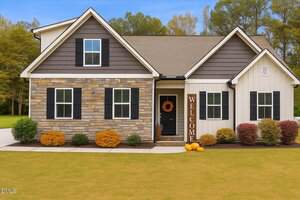 A one-story house at 4304 S Greg Allen Way with stone and white siding, black shutters, a black front door with an orange wreath, a "WELCOME" sign, three pumpkins on the porch, and shrubs along the front, set against a green lawn.