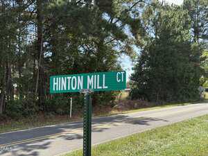 A green street sign reading "HINTON MILL CT" stands beside a two-lane road near 3718 Hinton Mill Court, Franklinton NC 27525, with grass and trees lining the way under a clear sky.