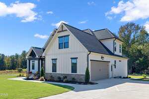 A modern two-story house with light-colored siding, dark roof shingles, black trim, and a two-car garage. The home sits on a manicured lawn with a concrete driveway and small landscaped garden beds.