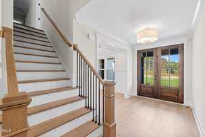 A bright entryway with light wood floors, a wooden staircase with black balusters, white walls, a large light fixture, and double front doors with glass panels, looking out to a green lawn.
