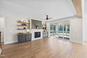 A modern living room with light wood floors, built-in shelves and cabinets flanking a white fireplace, a ceiling fan, and large sliding glass doors opening to a backyard with trees.