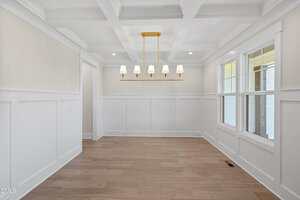 A bright, empty dining room with white wainscoting, a coffered ceiling, light wood flooring, large windows, and a gold chandelier with six lampshades hanging in the center.