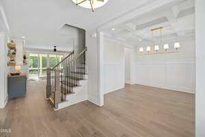 A bright, open interior features light wood floors, a staircase with dark railings, a dining area with a coffered ceiling and chandelier, and large windows leading to a patio with a view of greenery.