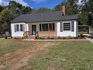 A single-story white house with black shutters, a covered front porch with wooden railings, a light blue door, a brick chimney, and a driveway leading to a carport on the right, surrounded by grass and trees.