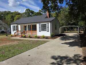 A small white house with black shutters, a front porch with wooden railings, and a concrete driveway leading to a covered carport. The yard is partially grassed with some landscaping near the house.