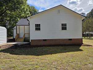 A white single-story house with brick foundation, horizontal siding, two windows, and a small porch with wooden steps. The yard is grassy with some trees and a partly cloudy sky overhead.