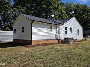 A white, single-story house with black trim and a gray roof is shown from the side. The exterior is vinyl siding, and there is an HVAC unit on the right. The house sits on a red brick foundation. Trees are in the background.