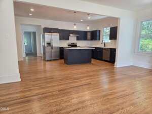 Open kitchen with dark cabinets, stainless steel appliances, center island, and white tile backsplash. Kitchen opens to a large room with hardwood floors. Multiple windows let in natural light.