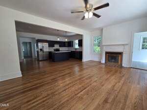 A spacious open-plan room with hardwood floors, a ceiling fan, a white fireplace with a brick interior, and a modern kitchen featuring dark cabinets and stainless steel appliances. Large windows provide natural light.