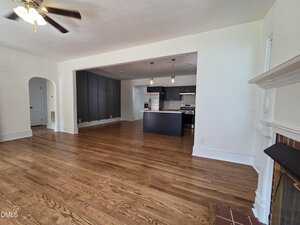 Open living space with hardwood floors, white walls, a ceiling fan, a fireplace on the right, and a modern kitchen with dark cabinets and stainless steel appliances in the background.