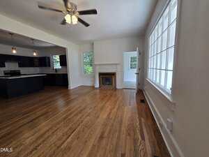 A bright living room with hardwood floors, a ceiling fan, a fireplace with a mirror above it, large windows on one wall, and an open view into a modern kitchen with dark cabinets and pendant lights.
