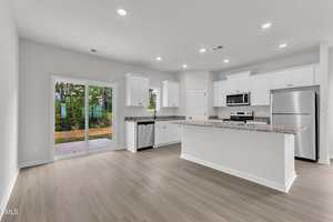 A modern kitchen with white cabinets, stainless steel appliances, granite countertops, and light wood flooring. Large sliding glass doors on the left open to a patio and wooded outdoor area. Recessed lighting is on the ceiling.