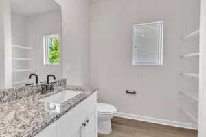 A bathroom with a granite countertop sink, black faucet, white cabinets, a toilet, open shelving, two small windows with blinds, and light wood flooring. The walls are painted white.