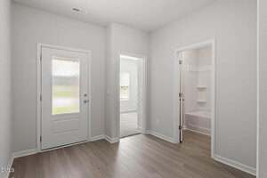A hallway with light wood flooring and white walls, featuring a white front door with a frosted glass window, and two open doorways leading to a carpeted room and a bathroom with a bathtub.
