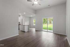 A bright, empty living area with wood floors, white walls, and a ceiling fan. Sliding glass doors lead to a green backyard. The open kitchen with white cabinets and recessed lighting is visible to the left.