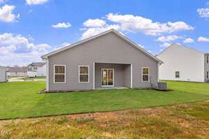 A single-story house with gray siding and a covered back patio, standing on a grassy lawn with an air conditioning unit on the right side. Other similar houses are visible in the background under a partly cloudy sky.