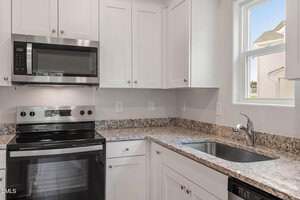 A modern kitchen with white cabinets, stainless steel appliances including a stove and microwave, granite countertops, and a single-basin sink beneath a small window.