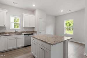 A modern, unfurnished kitchen with white cabinets, a granite-topped island, stainless steel dishwasher, and sink. Two windows and recessed lighting brighten the space. The flooring is light-colored wood.
