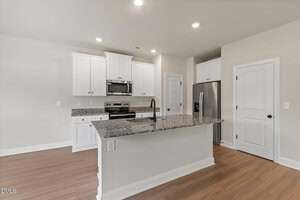Modern kitchen with white cabinets, stainless steel appliances, granite countertops, a center island with sink and faucet, and wood-style flooring. Walls are light-colored and lighting is recessed ceiling fixtures.