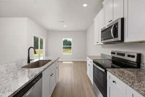 A modern kitchen with white cabinets, granite countertops, stainless steel appliances including a stove and microwave, a sink in the island, and light wood flooring. A window and glass door offer views of a grassy yard outside.
