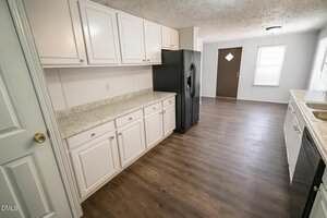 A kitchen with white cabinets, a black refrigerator, wood-look flooring, a marble-patterned countertop, a stainless steel double sink, and a brown door leading outside. Natural light enters through two windows.