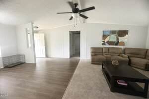 A living room with a ceiling fan, beige recliner sofa, dark wood coffee table, abstract wall art, and a section of carpeted and laminate flooring; a dog crate is near the front door.