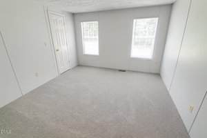 An empty room with light gray carpet, white walls, two windows with blinds, and a closed white door. Electrical outlets are visible on the walls, and natural light enters through the windows.