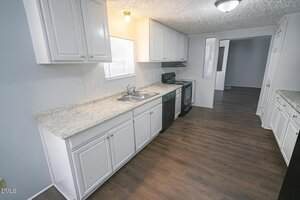 Galley kitchen with white cabinets, light countertops, stainless steel sink, dishwasher, stove, and wood-patterned flooring. A window above the sink provides natural light, and an open doorway leads to another room.