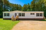 A single-story manufactured home at 141 Cardinal Lane with white siding, black shutters, a brick foundation, and two wooden porches, set on a grassy lawn with a gravel driveway and trees in the background.