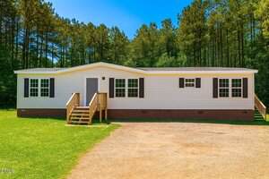 A single-story manufactured home at 141 Cardinal Lane with white siding, black shutters, a brick foundation, and two wooden porches, set on a grassy lawn with a gravel driveway and trees in the background.