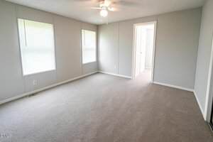 An empty room with light gray walls, beige carpet, two windows with blinds, a ceiling fan with lights, and an open doorway leading to another room.
