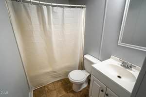 A small bathroom with light-colored walls, a white shower curtain, a white toilet, and a white vanity with a sink and faucet. A mirror hangs above the vanity. The floor is covered with tan tiles.