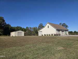 A single-story beige house with a brown roof sits on a large, grassy lawn at 8095 Crawford Currin Road. A detached beige garage or storage building is nearby, with trees lining the background under a clear blue sky.