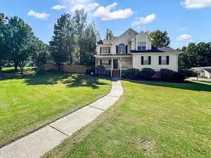 A two-story house with light-colored siding, a covered front porch, and an upper balcony, set on a large, well-maintained lawn with a curved concrete walkway leading to the front steps. Trees and shrubs surround the house.