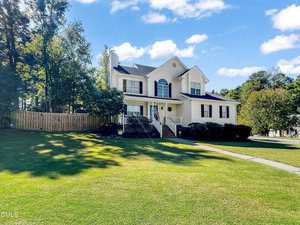 A two-story, light-colored suburban house at 2634 Bowden Drive with black shutters, a front porch, and steps leading to the entrance. The home is surrounded by a large green lawn, mature trees, and a wooden fence on one side.