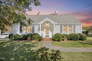 Single-story white house with gray roof, yellow window shutters, and a small covered front porch. The yard has trimmed bushes, potted plants, and a curved walkway. A car is partially visible to the left. The sky has a pink and blue gradient.