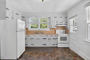 A kitchen with white cabinets and black hardware, a white refrigerator, stove, and sink under a window with blinds. There is a door and two windows on the right, and the floor has a brown tile pattern.